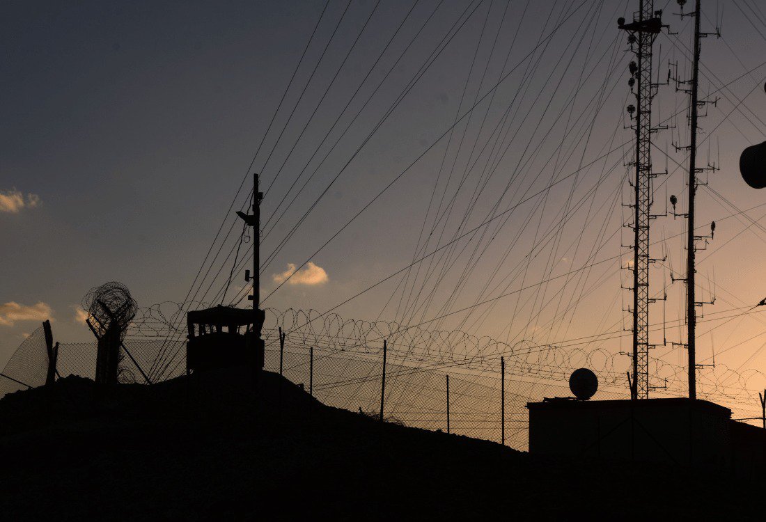 The sun sets behind a military communication tower, casting a warm glow over the landscape.